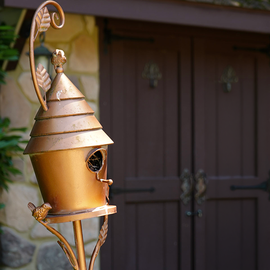 A decorative copper-toned metal birdhouse with a pointed roof, small bird perched on the stand, and a small bird perched on the front, photographed outdoors in front of dark brown double doors.