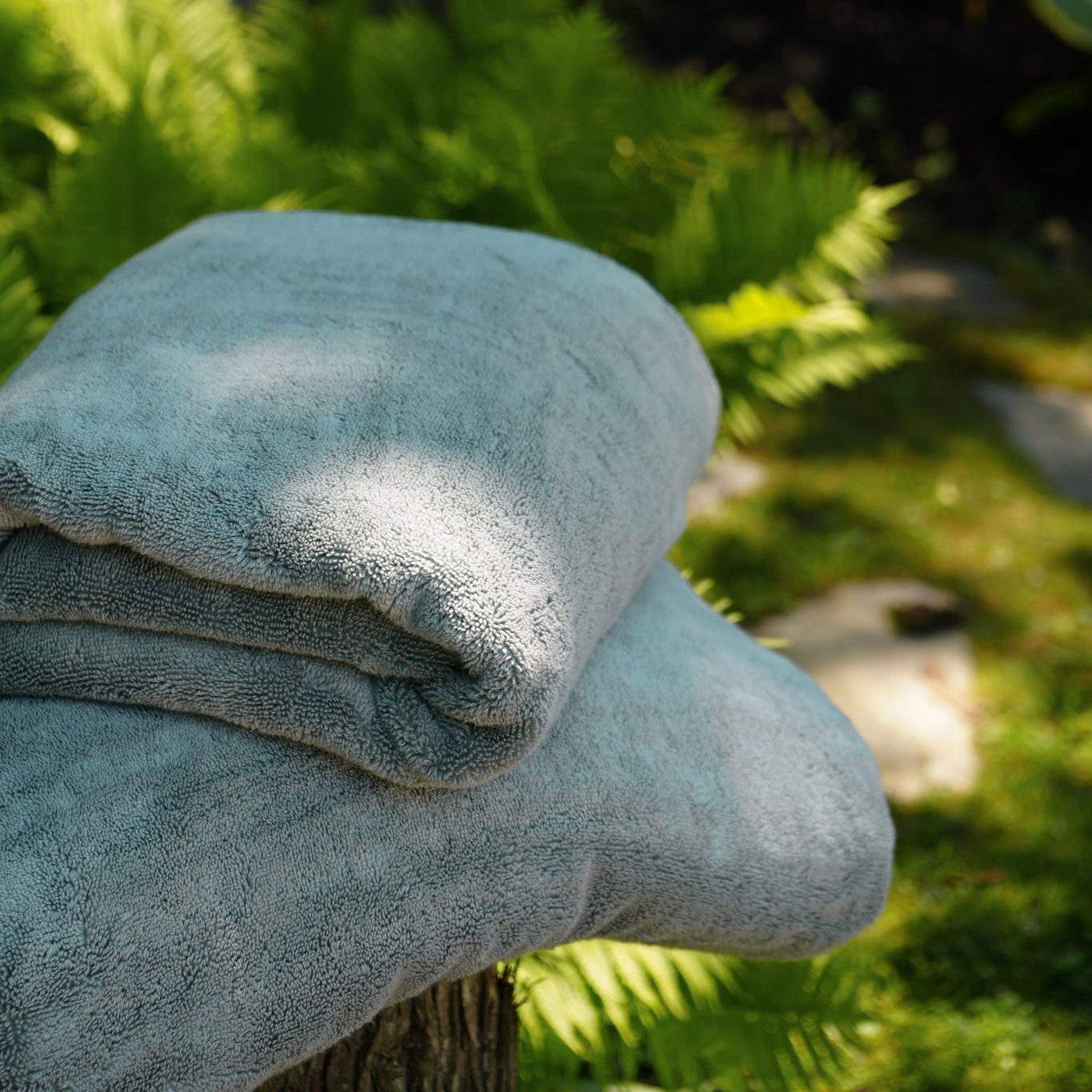 A folded light gray bath towel resting on a wooden surface outdoors, with green ferns and a stone wall blurred in the background.