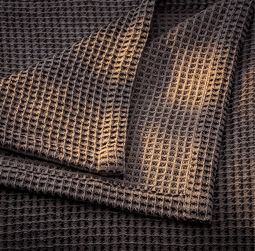 A close-up of a dark charcoal gray waffle-weave fabric, folded to show its textured grid pattern under warm directional light.
