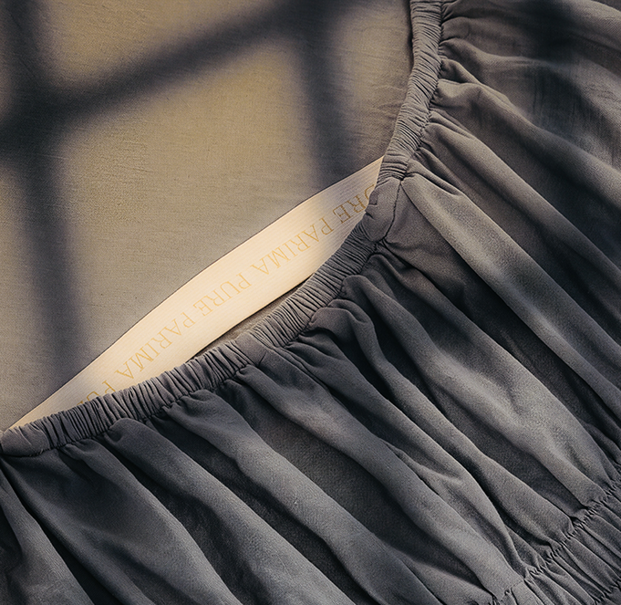 Close-up of a dark gray pleated fabric garment with elasticized gathered edges and a cream inner label reading 'PURE PARIMA'