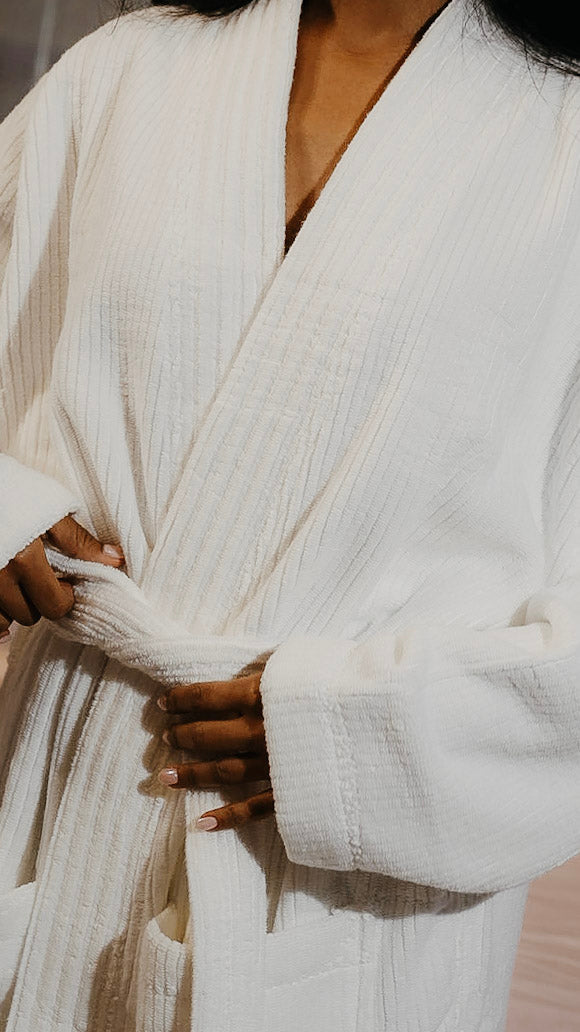 A close-up of a person wearing a white textured cotton robe, tying the belt at the waist. The robe has a deep V-neck and wide cuffs.