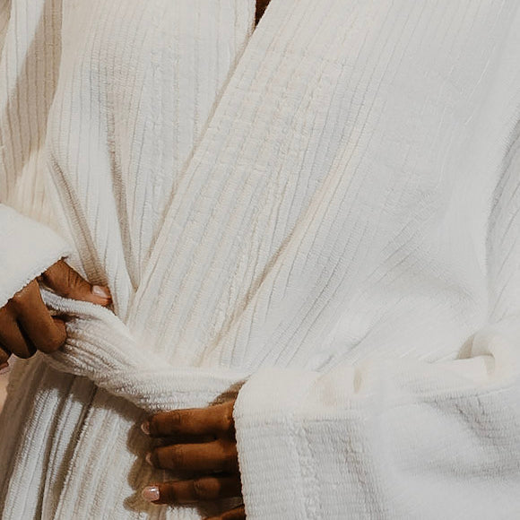 A close-up of a person wearing a white textured cotton robe, tying the belt at the waist. The robe has a deep V-neck and wide cuffs.