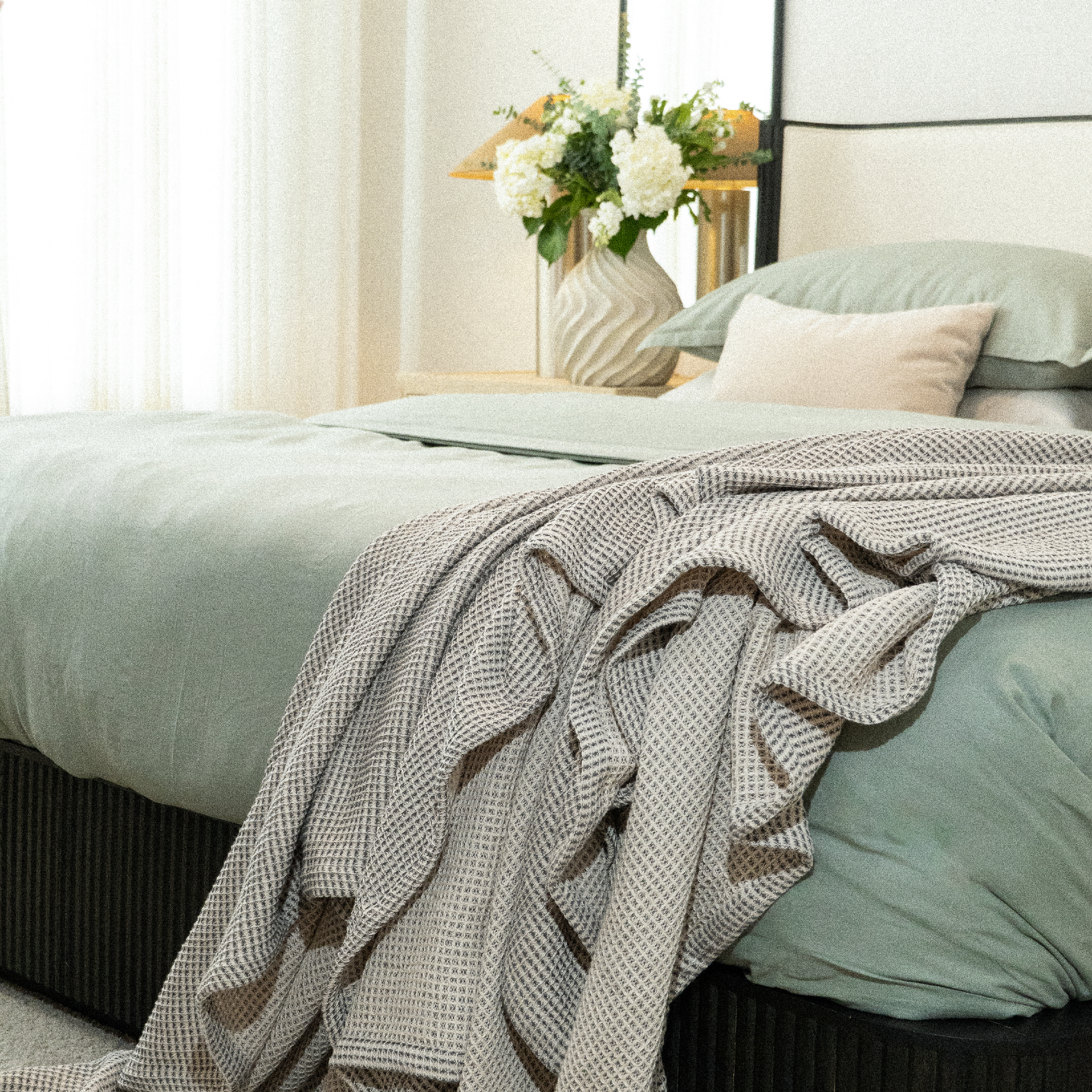 A styled bedroom scene showing a bed with sage green bedding and a textured beige-and-gray patterned throw blanket draped over the foot of the bed. A vase of white flowers and a mirror are visible in the softly lit room.