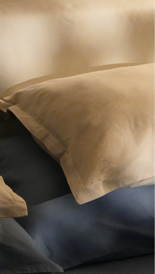 A close-up of a beige pillowcase resting on a dark blue bedsheet or duvet, with a softly blurred warm-toned background.