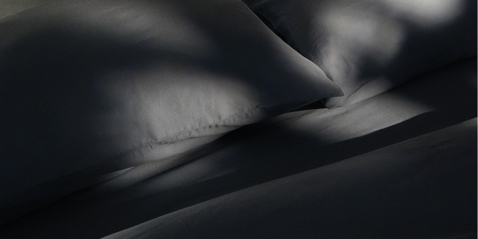 Dark charcoal gray bedding, including a pillow and sheets, shown in dramatic, high-contrast sunlight and shadow.