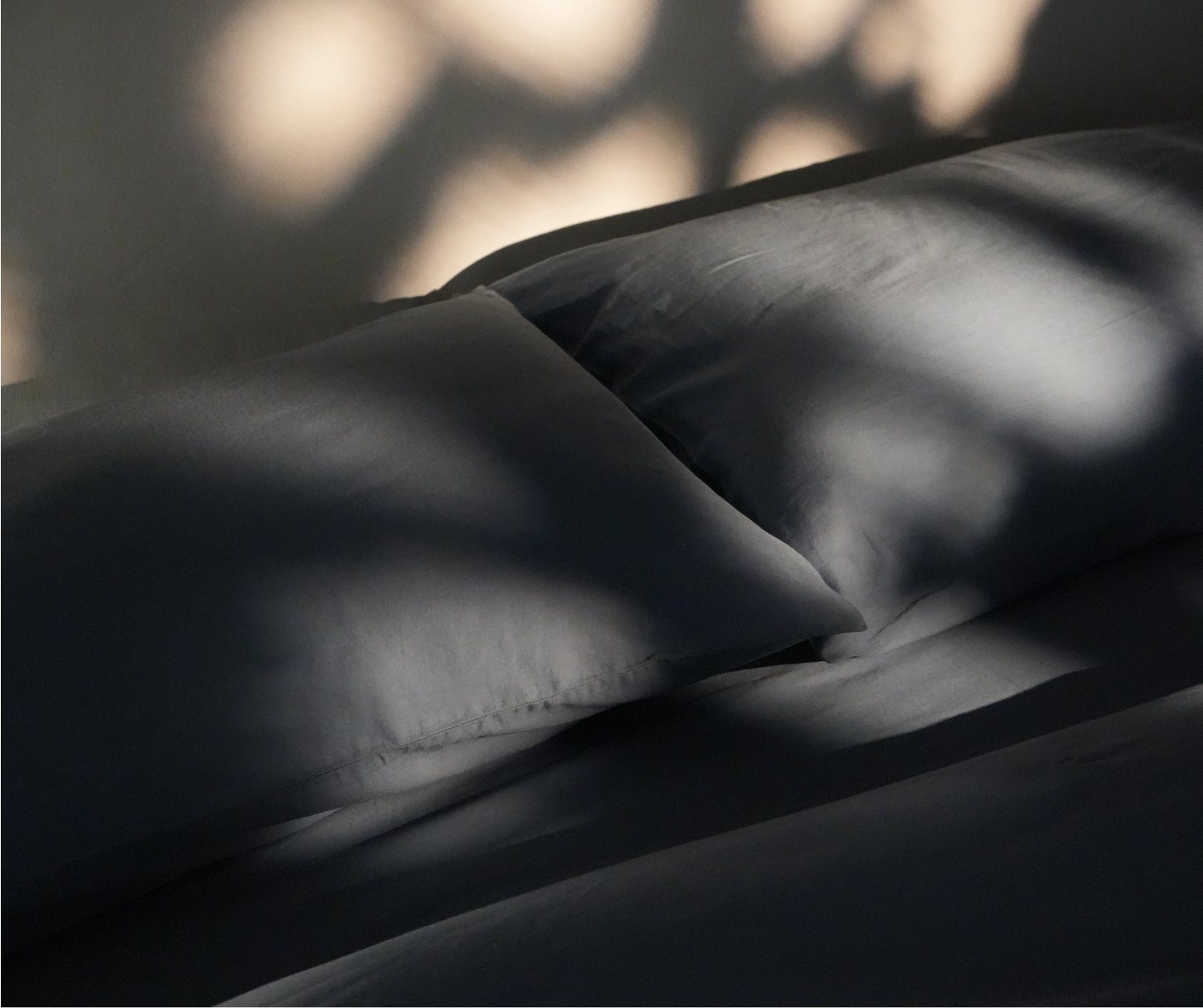 Dusky blue bedding, including pillows and sheets, styled with dramatic natural light and shadow patterns.
