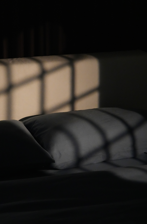 A dimly lit bed with pillows and bedding, featuring dramatic window-grid shadows cast across the surface in a moody interior scene.