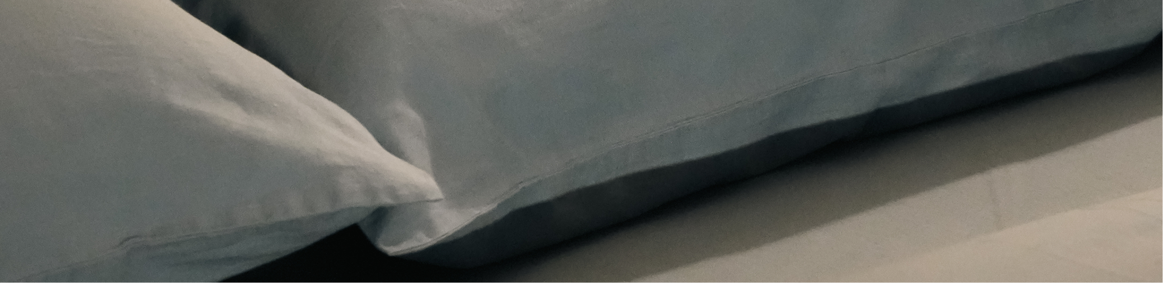 A close-up of a light blue or gray pillowcase with a white sheet underneath, showing the fabric texture and fold.