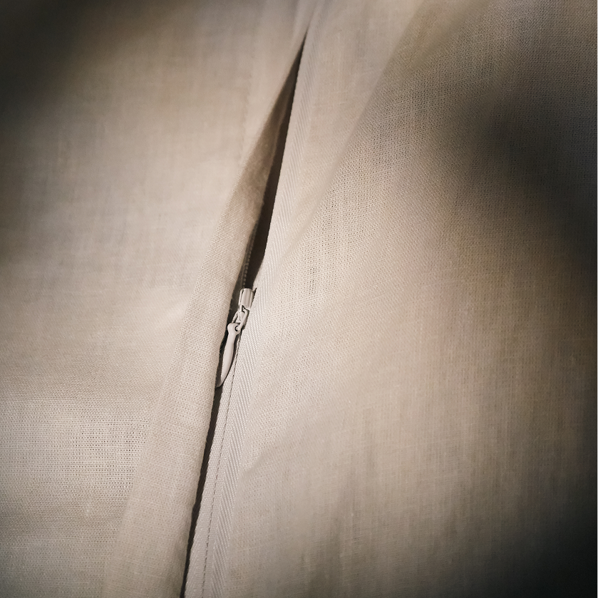 Close-up of a light beige fabric garment with a central zipper running vertically, showing the textured material and zipper pull in warm neutral tones.