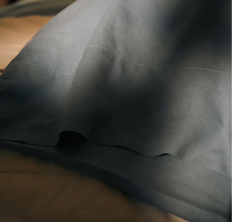 A close-up of a dark gray pillowcase with visible white stitching, resting on a bed with a beige duvet or sheet underneath.