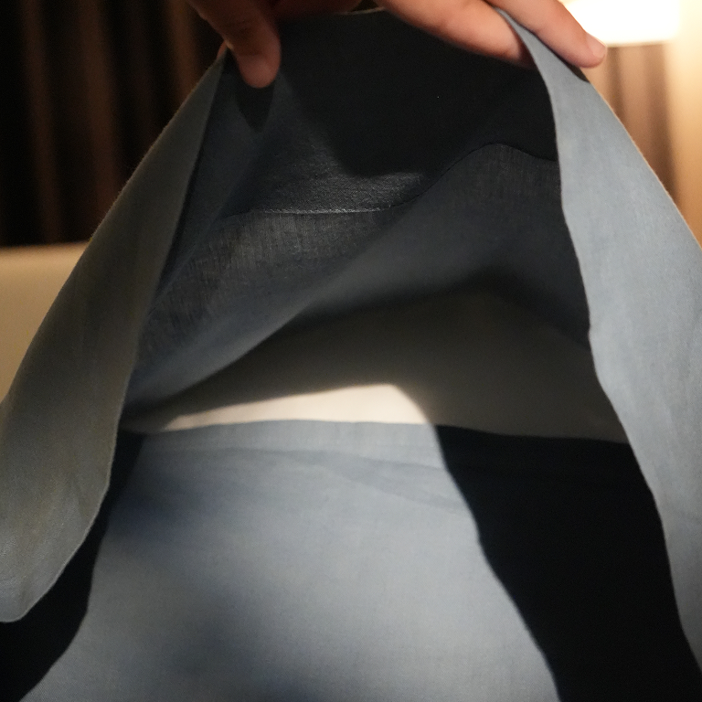 A close-up of a person holding open a light blue-gray fabric garment or bag, showing the interior lining and texture. A hand is visible at the top, and the background is a softly lit indoor setting.