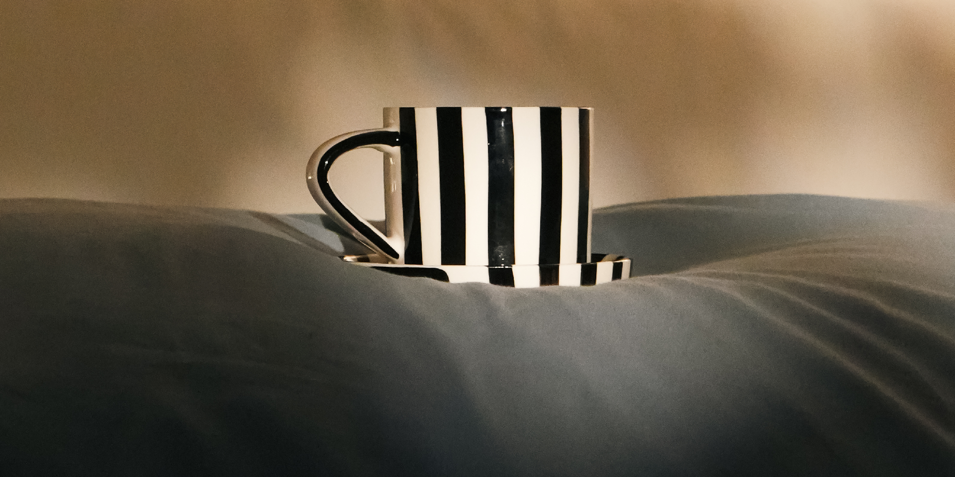 A black and white vertically striped ceramic mug with a matching saucer, resting on a dark gray pillow in a dimly lit room.