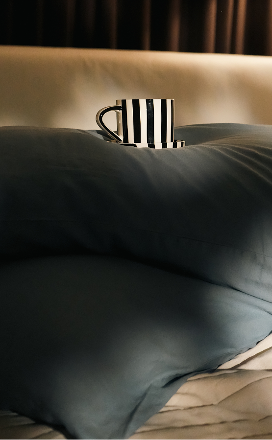 A black and white vertically striped ceramic mug sits on a dark slate blue pillow on a bed with beige bedding and a dark curtain in the background.