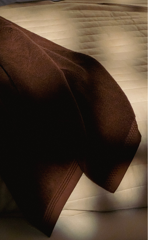 A close-up of a dark brown textured fabric, likely a towel or blanket, draped over a light beige quilted surface with warm, directional lighting.