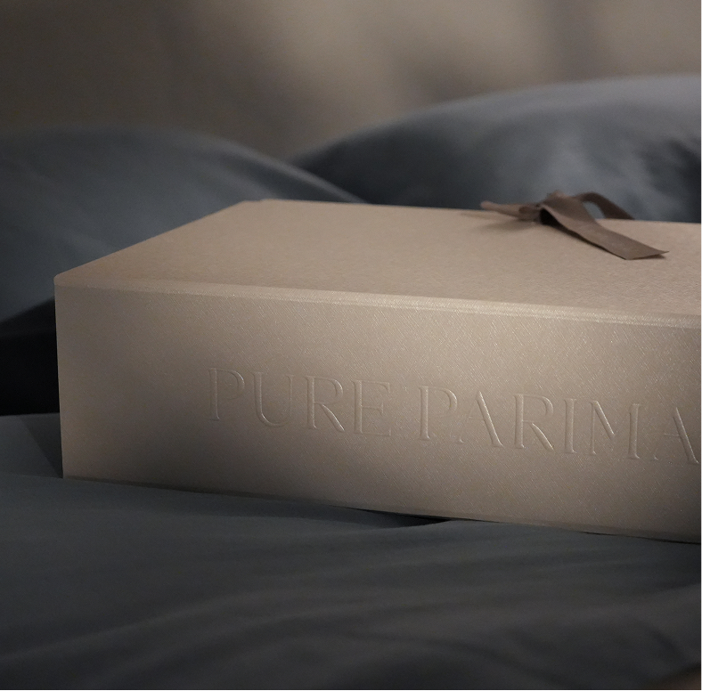 A beige textured box with the embossed brand name 'PURE PARIMA' on the side, tied with a brown ribbon, resting on dark gray bedding.