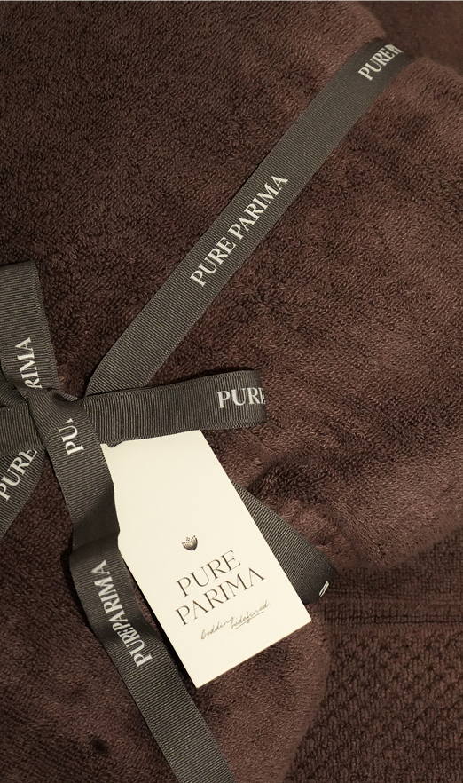 A close-up of a dark brown textured towel tied with a dark gray ribbon and attached white paper tag, both printed with the brand name PURE PARMA.