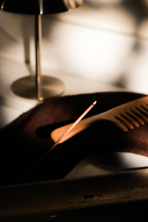 A close-up, warm-lit scene showing a dark brown leather hair tool or accessory with a comb-like edge and a thin metallic rod resting across it, alongside a wooden comb. A lamp base is blurred in the background.