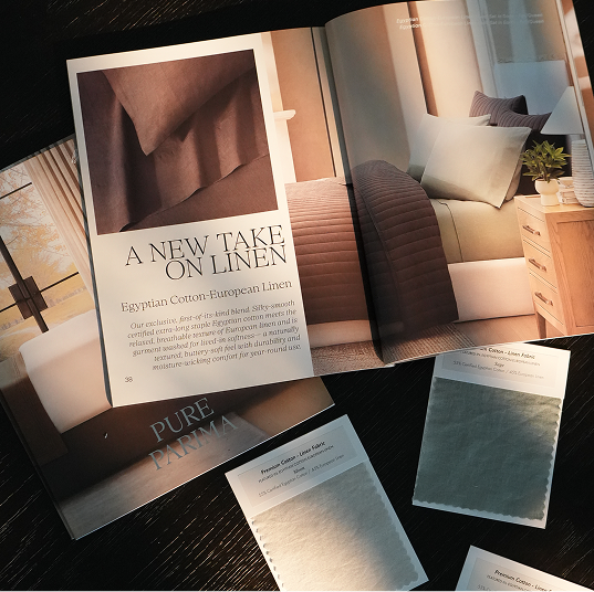 An open catalog or brochure spread for Pure Parma linen linens, showing a bedroom scene with bedding, a fabric swatch card, and printed sample sheets on a dark wooden surface.