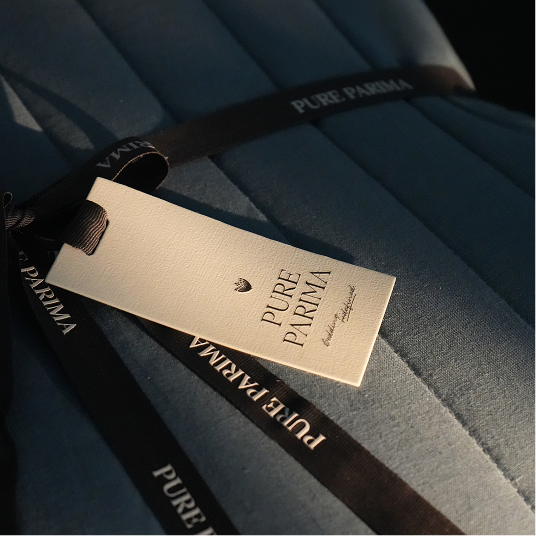 A close-up of a blue quilt with a cream paper tag and black ribbon printed with the brand name PURE PARMA.