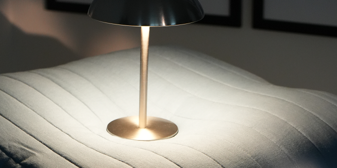 A modern table lamp with a dark metal dome shade and a warm brass-colored stem and base, casting a soft glow onto a white quilted surface.