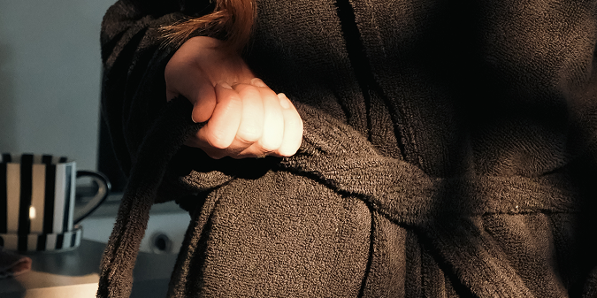 A close-up of a person wearing a black textured bathrobe, pulling the belt to tighten around the waist. A striped mug is visible in the background on the left.