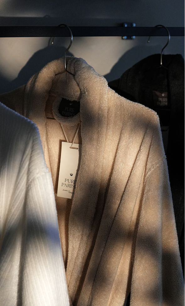 A beige plush bathrobe hanging on a black clothing rack, with a white tag reading 'PURE PARIS' visible. Strong diagonal sunlight casts deep shadows across the robe, and another white robe is partially visible in the foreground.