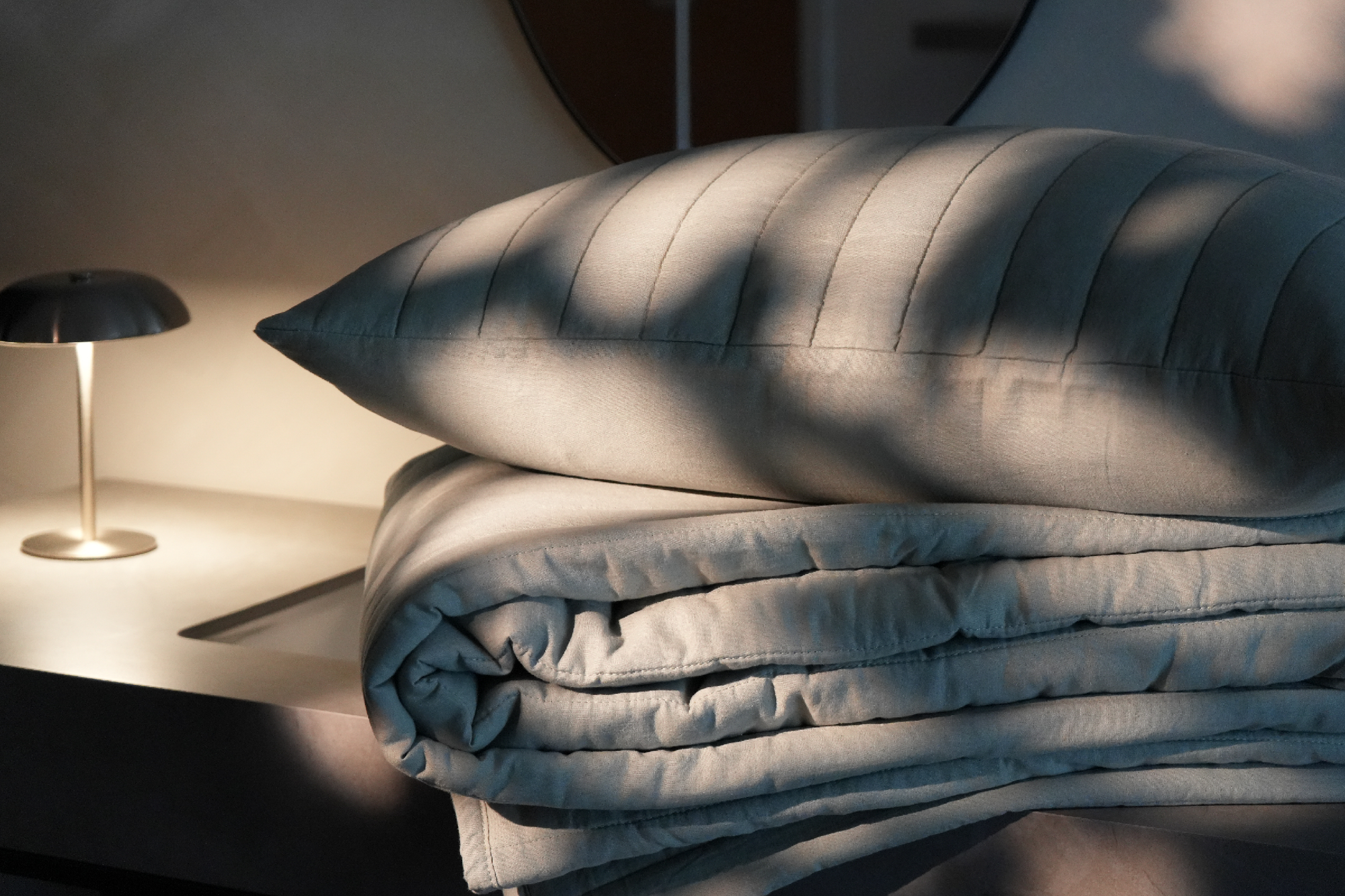 A light gray quilted pillow and matching folded bedding set in a softly lit bedroom scene with warm sunlight and shadows.