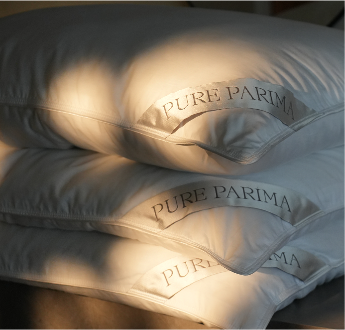 Three white fluffy pillows stacked together, each with a stitched label reading 'PURE PARIMA' in dark lettering, photographed in warm direct sunlight with strong shadows.