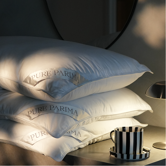 Three stacked white pillows with 'PURE PARIMA' embroidered on them, styled on a bedside table next to a striped mug in warm, moody lighting.