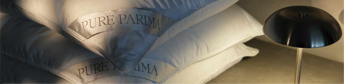 A stack of white pillows with 'PURE PARIMA' embroidered on them, shown next to a black dome-shaped table lamp in a dimly lit room.