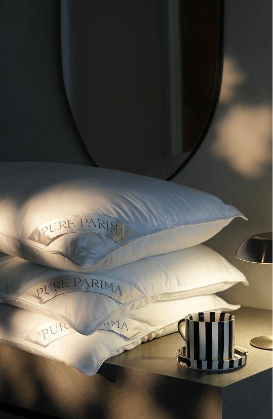 Three stacked white Pure Parima pillows on a bedside table, lit by warm sunlight with a striped mug and a lamp nearby.