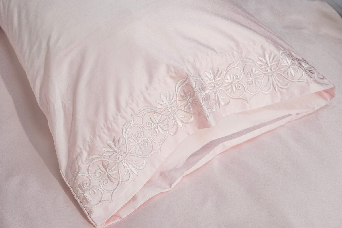 A close-up of a light pink pillowcase with delicate scroll embroidery along the border, resting on matching pink bedding.