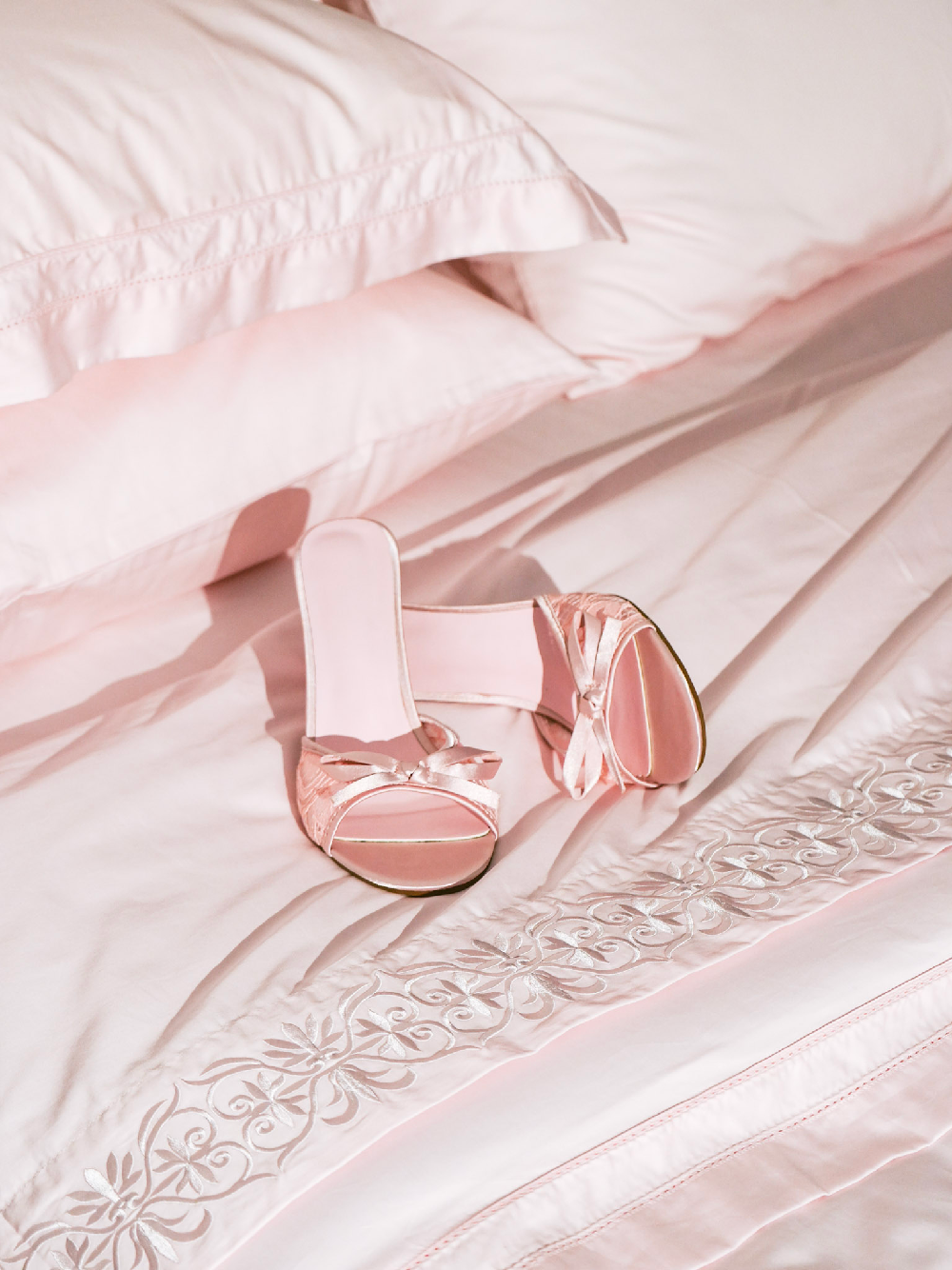 A pair of light pink satin mule sandals with bow details, placed on a matching pink embroidered bedsheet next to a pillow.