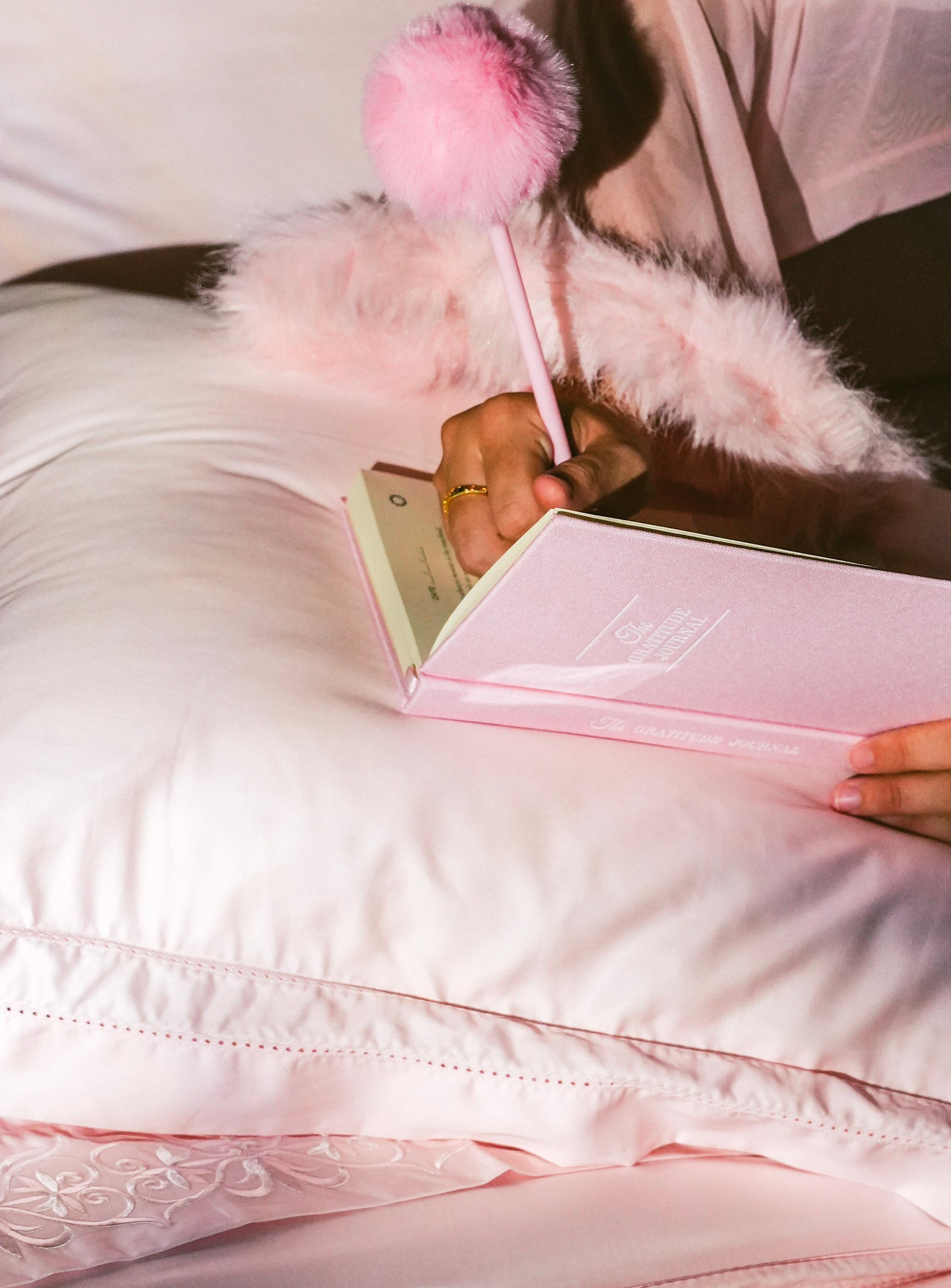 A person lying on a white bed, writing in a pink notebook with a pink pom-pom pen. The scene is softly lit in pastel pink tones, with fluffy bedding and a pink garment visible.