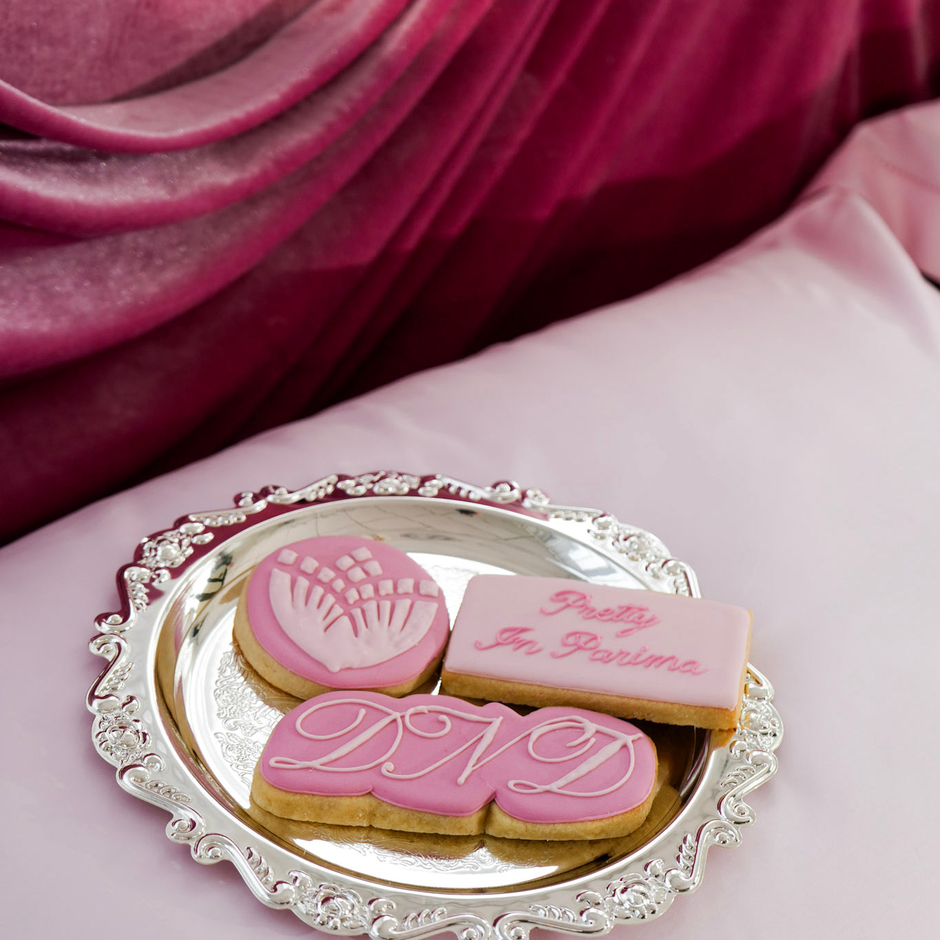 Three decorated sugar cookies on an ornate silver tray, set on a pink sateen surface with draped magenta fabric in the background. 