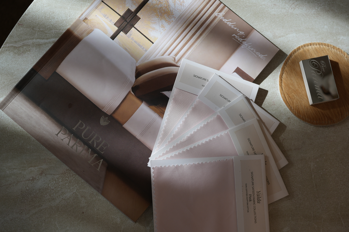 A styled flat-lay of a Pure Parima bedding color palette, featuring printed color swatches, a color wheel, and a color story booklet on a textured beige surface with soft natural shadows.