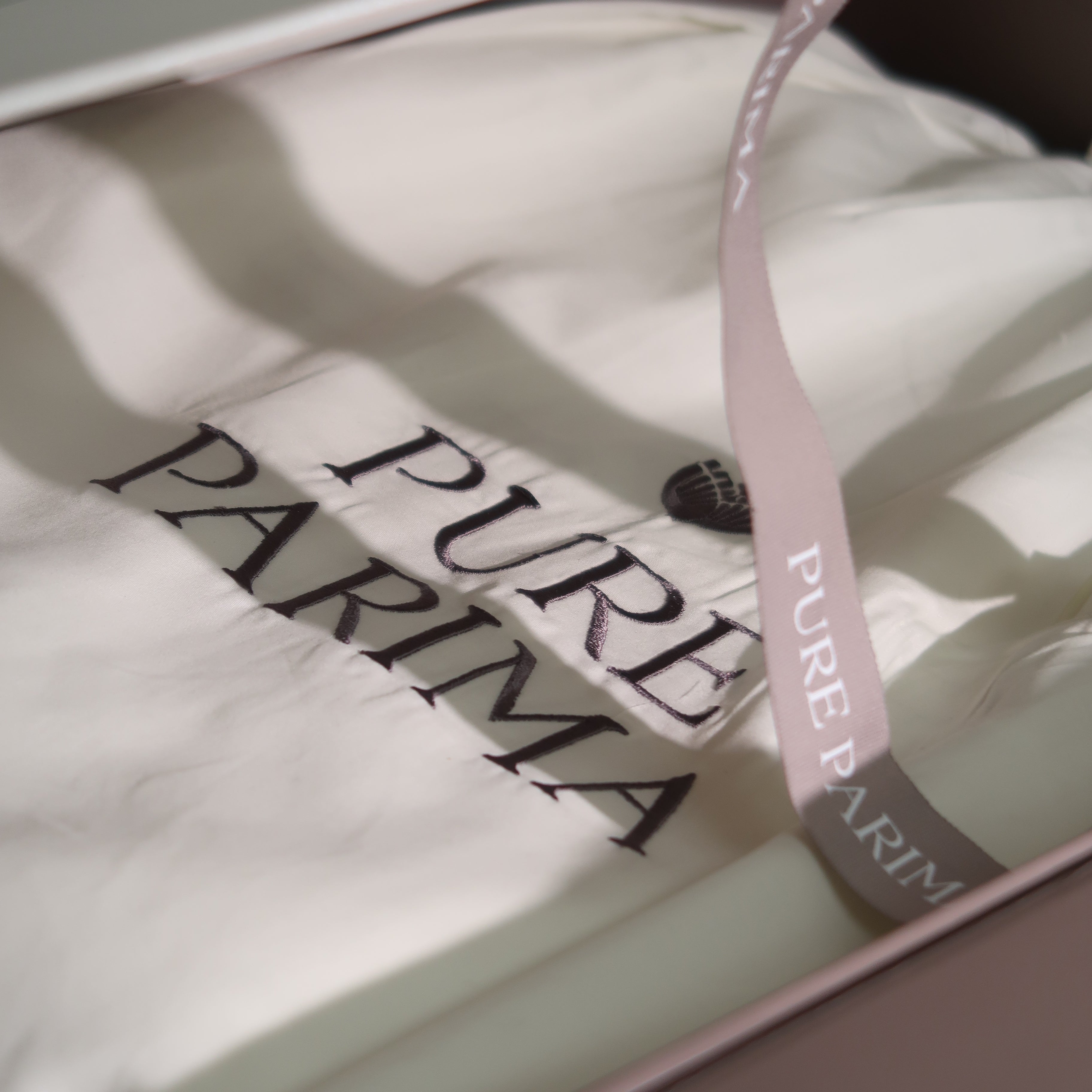 A folded cream-colored garment or fabric item inside a pink box, printed with the words 'PURE PARIMA'. A pale pink ribbon with repeating 'PURE PARIMA' text drapes across the top.