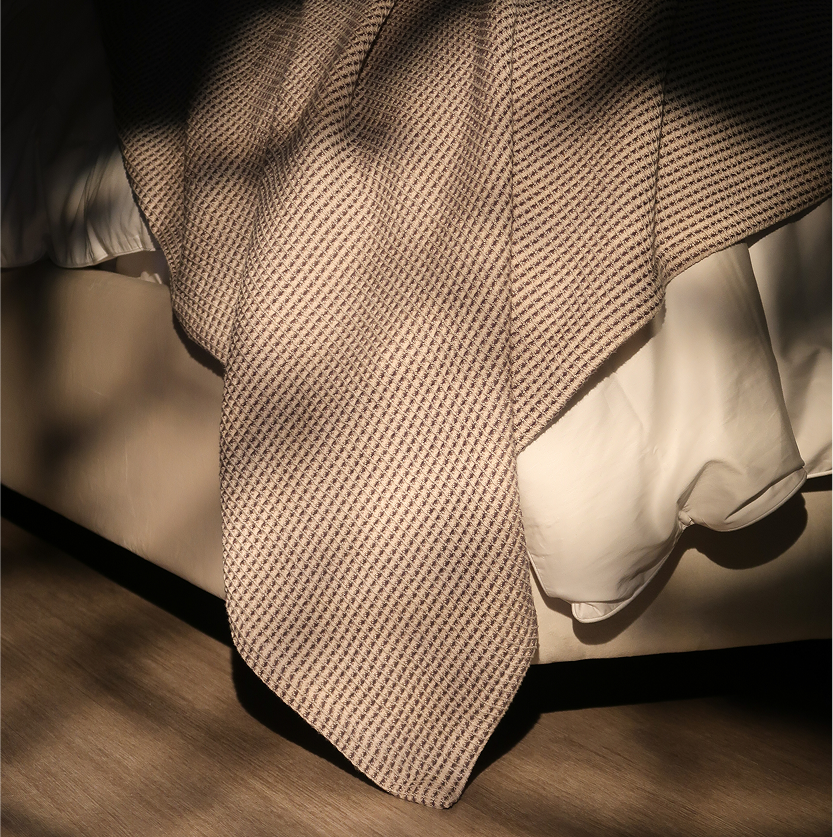 A close-up of a textured beige knit tie draped over white dress shirts on a bed, with warm sunlight casting leaf shadows across the fabric.