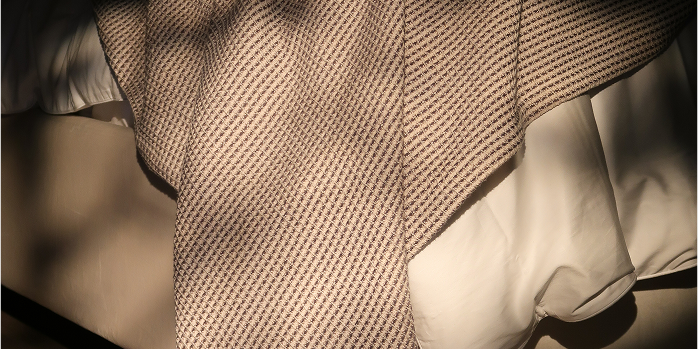 A close-up of a textured beige and brown woven fabric, likely a scarf or shawl, draped over a white garment with dramatic shadow patterns.