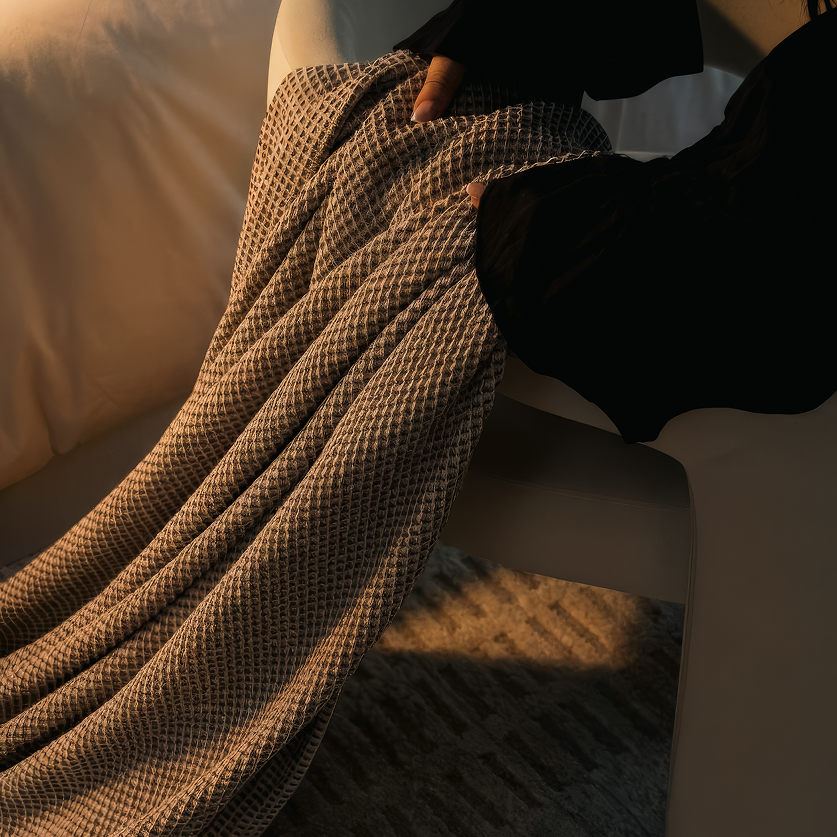 A close-up of a textured knit blanket in beige and black draped over a white chair in a warmly lit room.