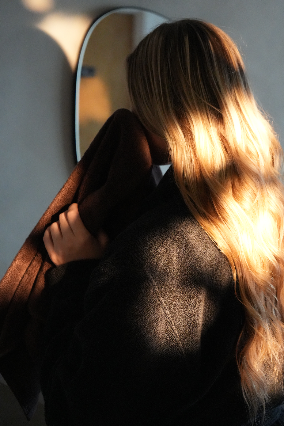 A woman with long blonde hair seen from behind, wrapped in a dark brown blanket or coat, standing in front of a mirror in dramatic, high-contrast sunlight and shadow.