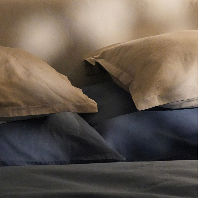 A close-up of a bed with neutral beige pillows and bedding, layered over dark blue and slate gray sheets, set against a dark brown headboard and curtains.