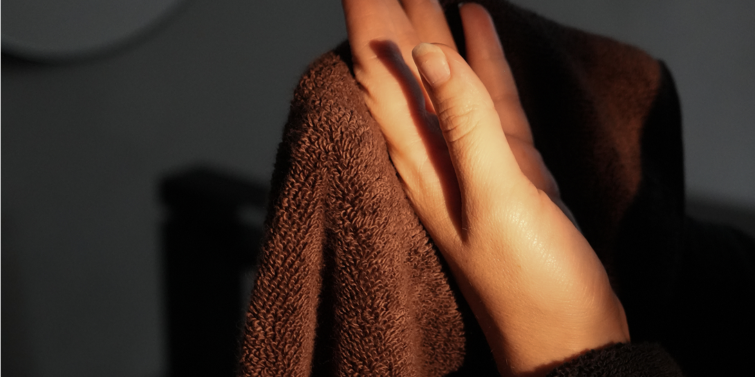 A close-up of a person's hand drying themselves with a dark brown textured towel in warm, low-key lighting.