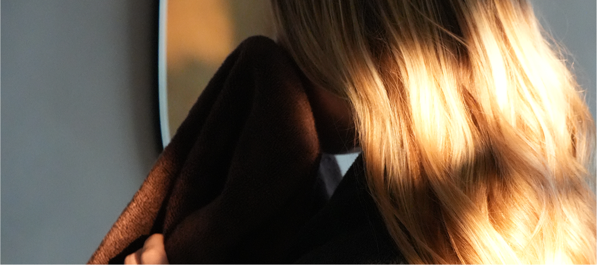 A close-up lifestyle shot of a person with long, wavy blonde hair wearing a dark brown shawl draped over their shoulders, with warm sunlight filtering through.