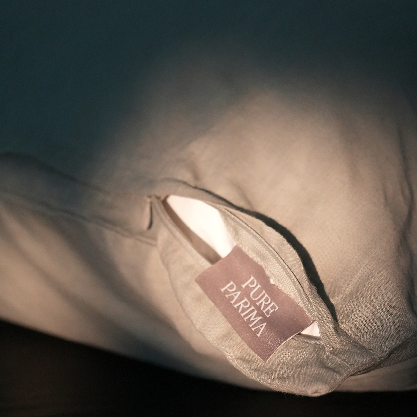 A close-up of a beige fabric pillow or cushion with a visible sewn label reading PURE PARMA, photographed in dim warm lighting against a dark background.