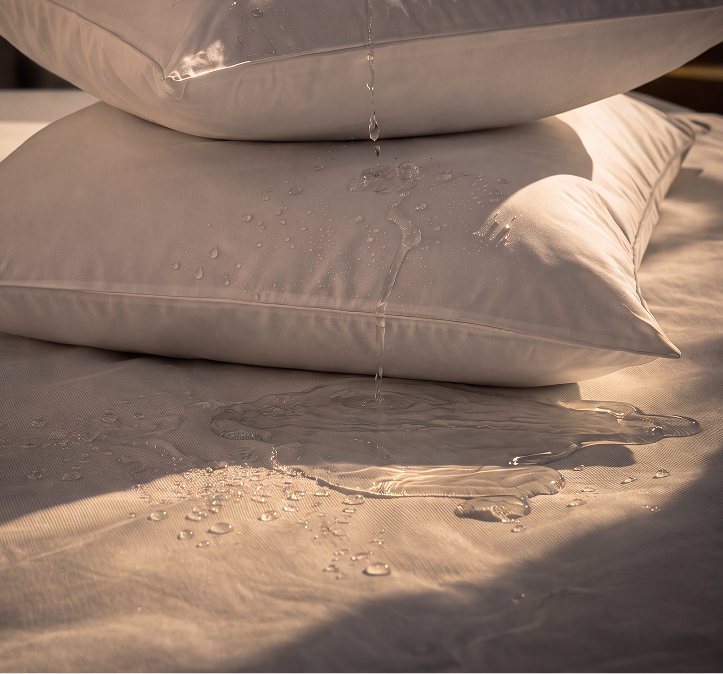 Two white pillows stacked on a bed with water droplets dripping from them onto a wet beige bedding surface, demonstrating water resistance.