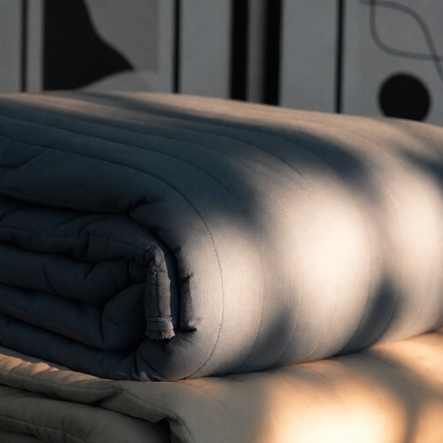 Two folded bedding items, likely a dark blue-gray quilt and a light beige duvet or pillowcase, stacked on a wooden surface with dramatic natural light and shadow patterns.
